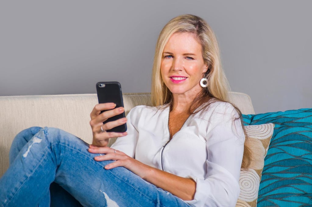 Eine blonde heiße Milf in weißer Bluse und Jeans sitzt entspannt auf einem Sofa, hält ein Smartphone in der Hand und lächelt in die Kamera.