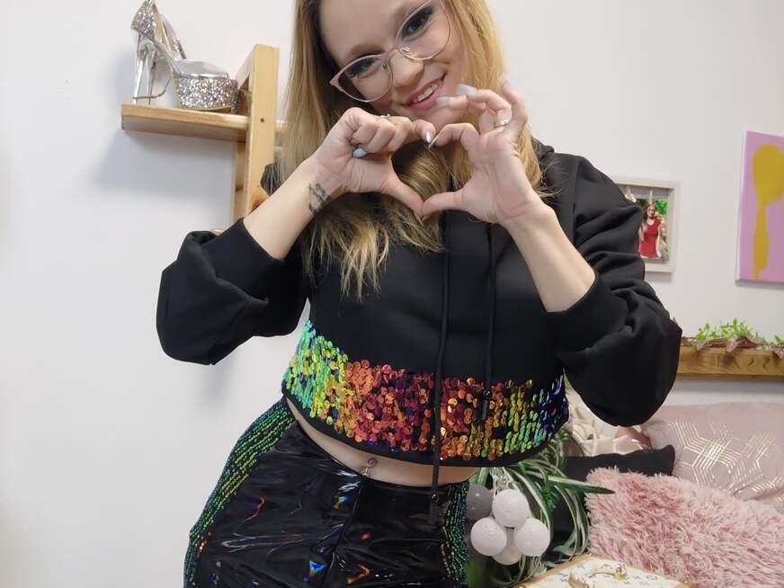 LeniFuchs in einem schwarzen Hoodie mit bunten Pailletten, formt ein Herz mit den Händen und lächelt verführerisch in die Kamera.