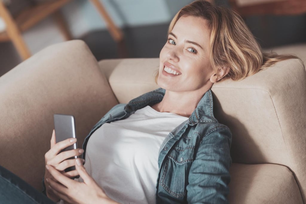 Eine lächelnde Sexy Milf mit blondem Haar, weißem T-Shirt und Jeansjacke liegt entspannt auf einem beigen Sofa und hält ein Smartphone in der Hand.