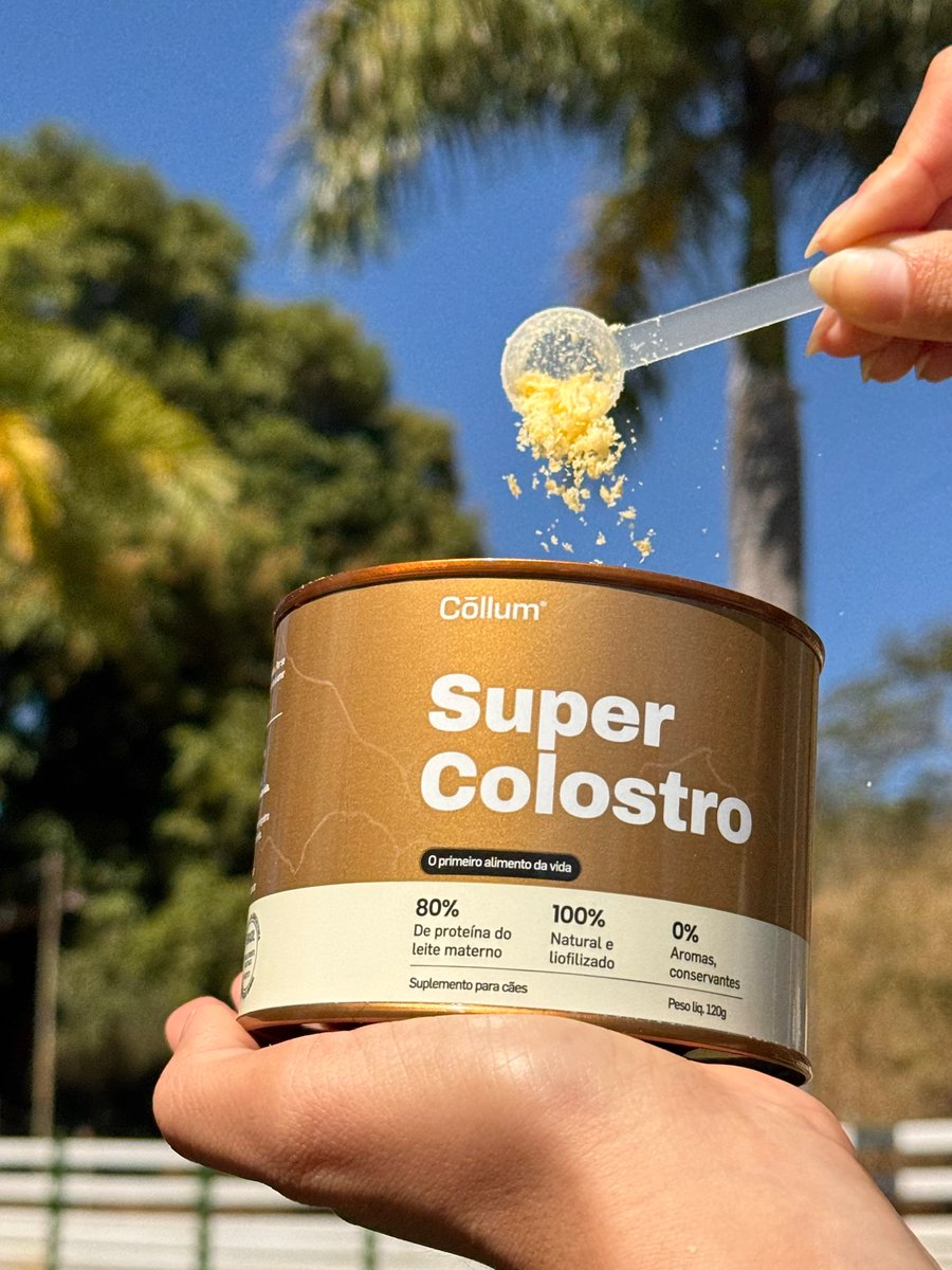 Mão servindo um scoop de Super Colostro no pote, com a fazenda ao fundo