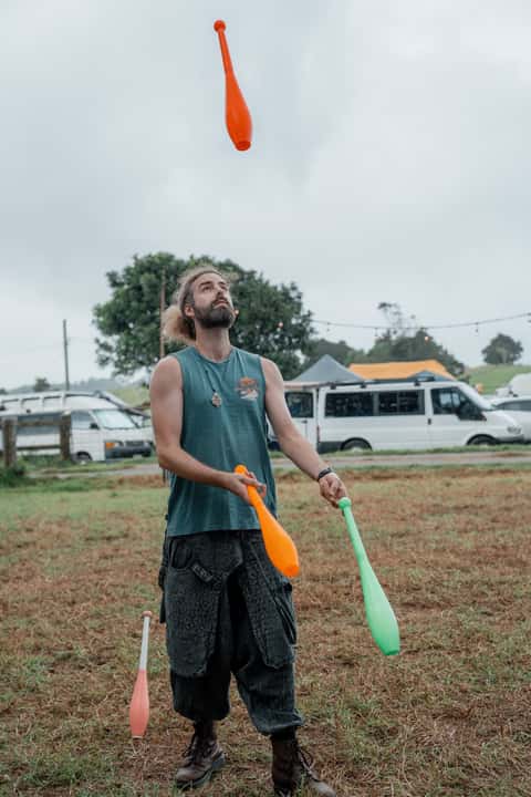 Juggler tossing orange and green clubs above his head at a daytime bush festival