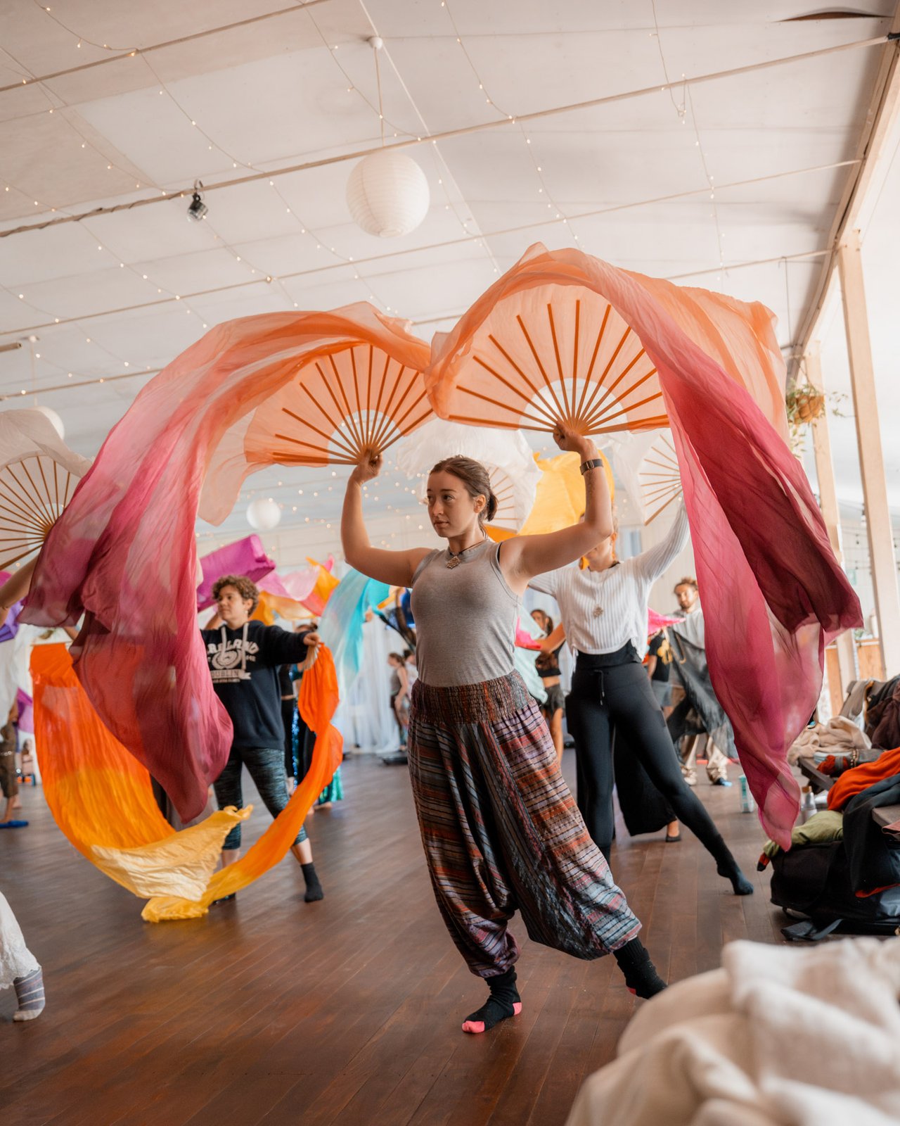 Two fan dancers spinning rose and coral fans inside a fairy-lit workshop room