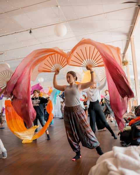 Two fan dancers spinning rose and coral fans inside a fairy-lit workshop room