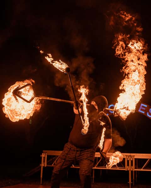 Fire performer mid-routine with double flaming fire-fan apparatuses at Glow Festival, night dark with ember field