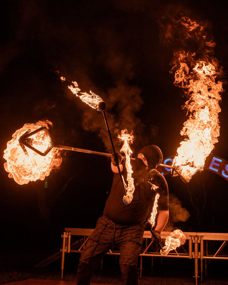 Fire performer mid-routine with double flaming fire-fan apparatuses at Glow Festival, night dark with ember field