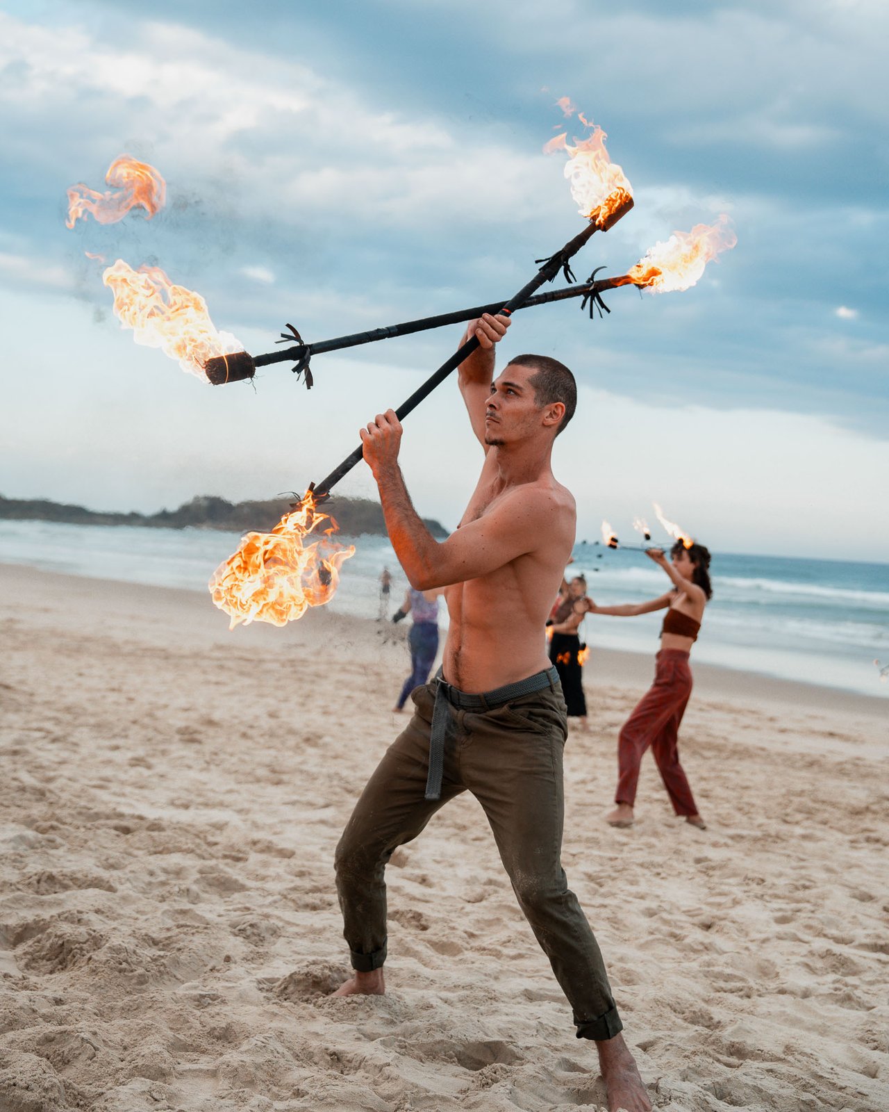 Fire performer spinning a flame staff on the beach at golden hour with the troupe in soft focus behind