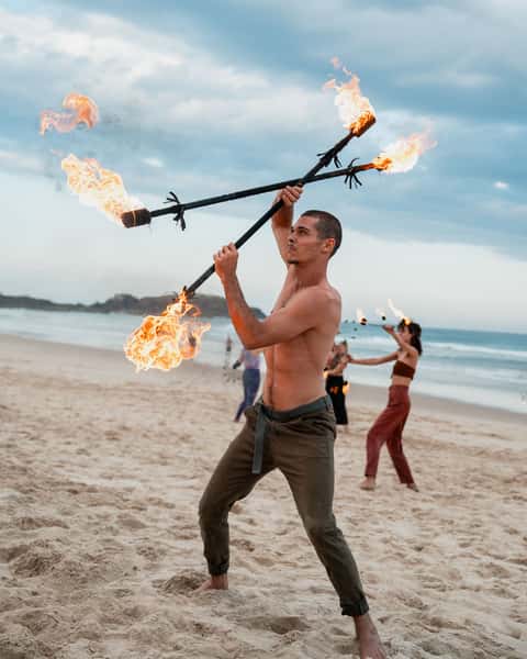 Fire performer spinning a flame staff on the beach at golden hour with the troupe in soft focus behind