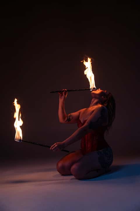Studio fire performer kneeling with two flame batons, red corset, controlled studio dark