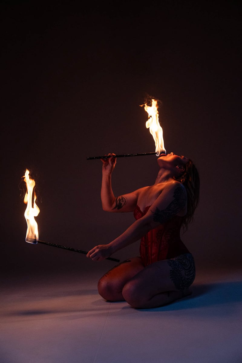 Studio fire performer kneeling with two flame batons, red corset, controlled studio dark