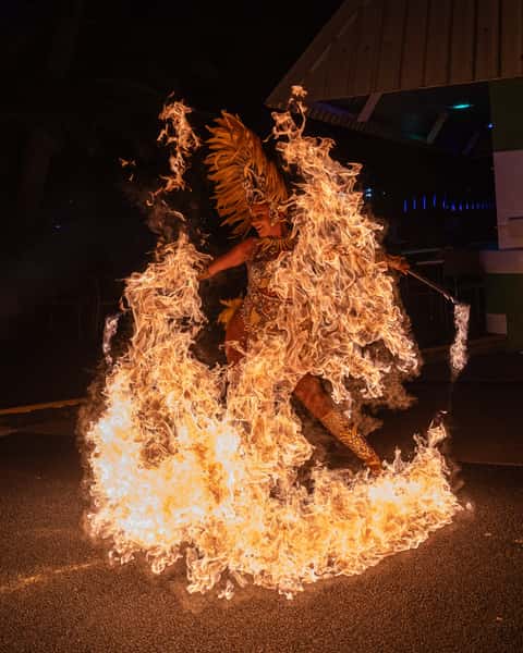 Performer in feathered headdress fully engulfed in a wash of flame on a dark road