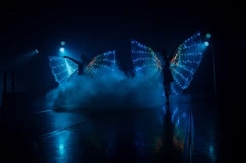 Two LED butterfly-wing performers on a smoky stage in deep blue beams
