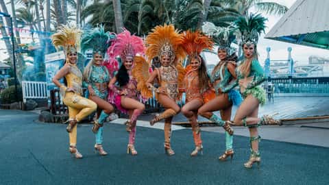 Samba dance troupe in oversized feathered headdresses lined up outside under daylight