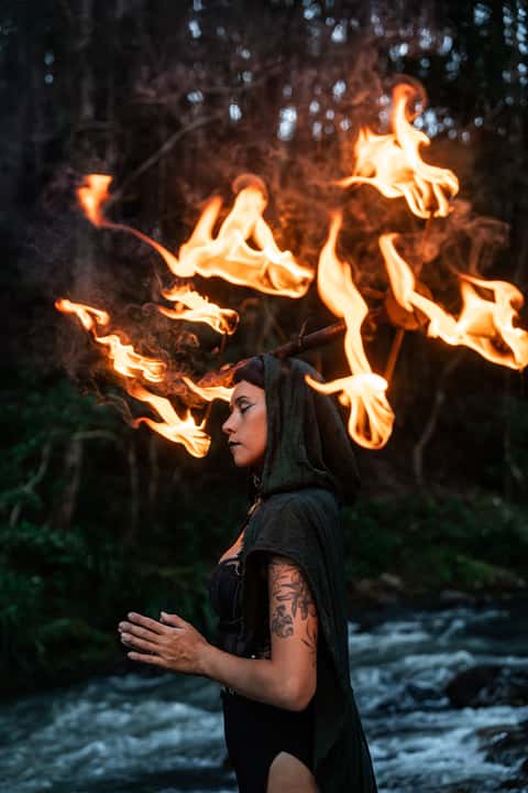 Fire performer in green hood with a halo of flame standing by a forest stream at dusk