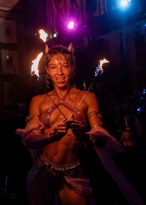 Fire performer with fire wands and crystal-fan headdress in a dark venue under purple and orange spot
