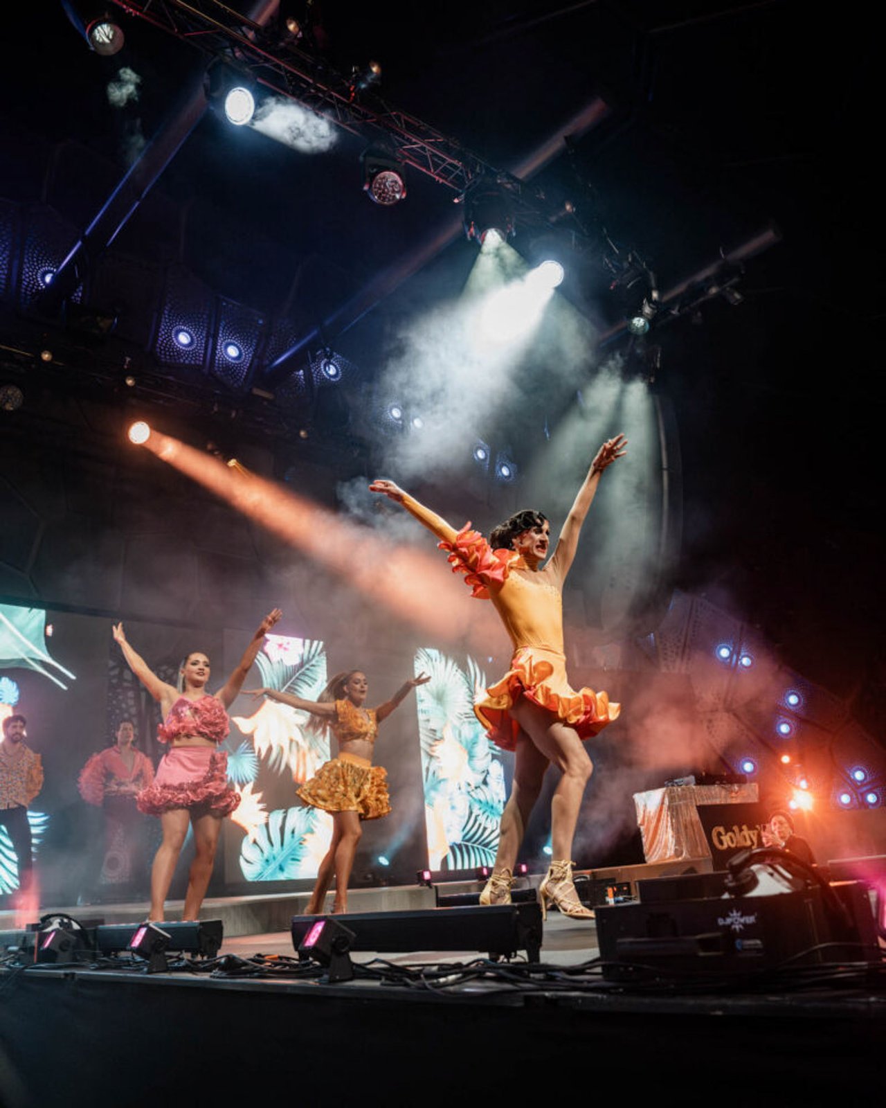 Latin dance troupe on main festival stage with spotlight and stage haze, costumed in ruffled coral and yellow