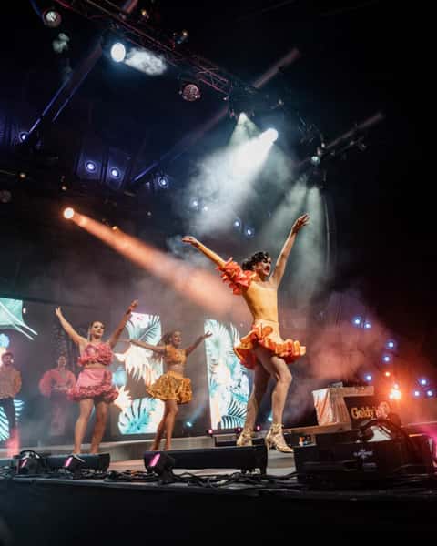 Latin dance troupe on main festival stage with spotlight and stage haze, costumed in ruffled coral and yellow