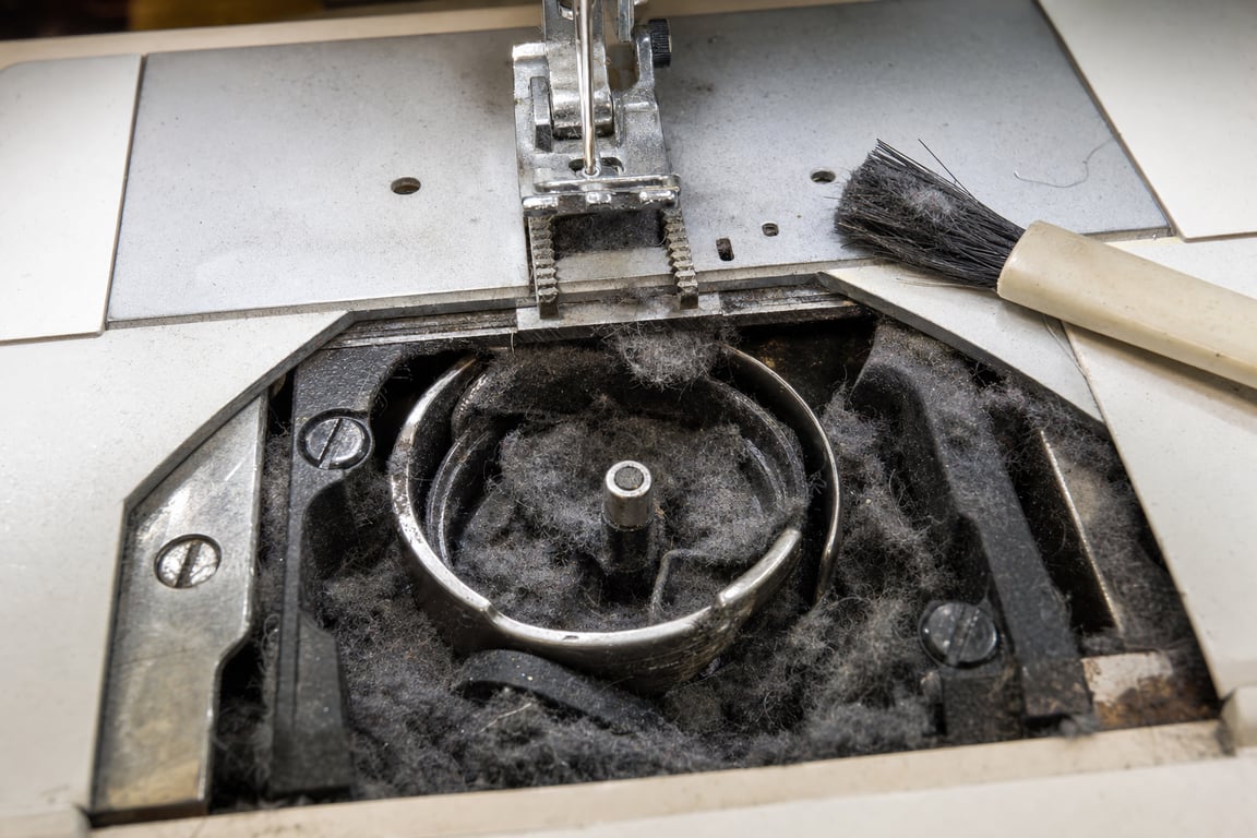 Close-up of a sewing machine's open bobbin area with feed dogs packed with gray felted lint, next to a small cleaning brush