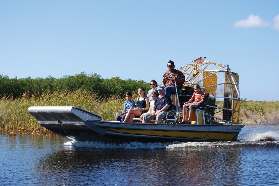 Airboat carrying a family and guide glides across Everglades water past tall sawgrass under a blue sky