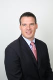 Smiling man in a dark suit, light blue shirt and pink tie posing for a studio headshot against a white background