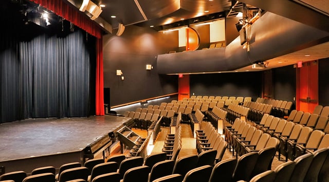Empty theater auditorium at TheatreZone with tiered rows of seats facing a bare stage framed by red curtains