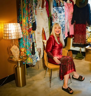 Smiling blonde woman in a red velvet dress seated in a gold chair amid colorful hanging clothing at Elsie In Naples