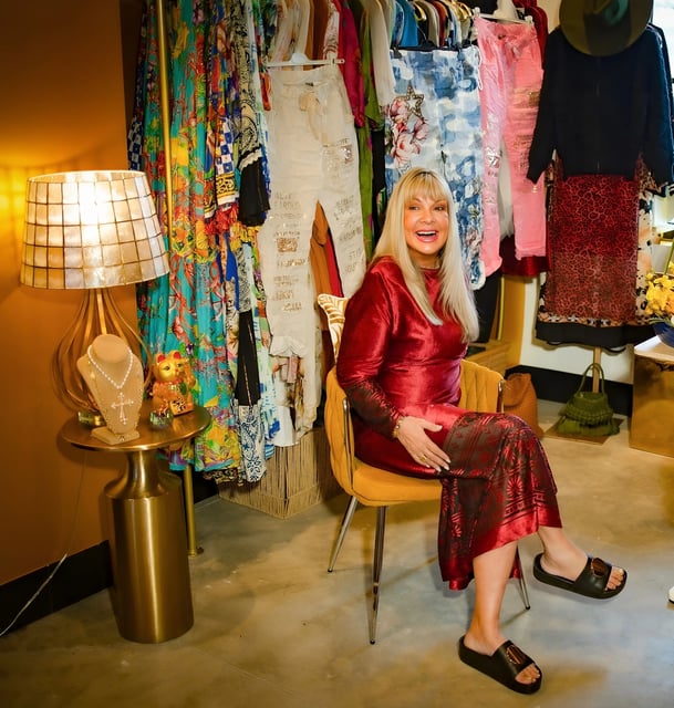 Smiling blonde woman in a red velvet dress seated in a gold chair amid colorful hanging clothing at Elsie In Naples