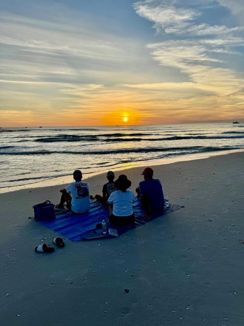 Four people sit on a blue blanket on the sand watching the sun set over the Gulf