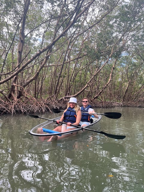 Two paddlers in life vests glide through a mangrove tunnel in a clear-bottom tandem kayak on calm water