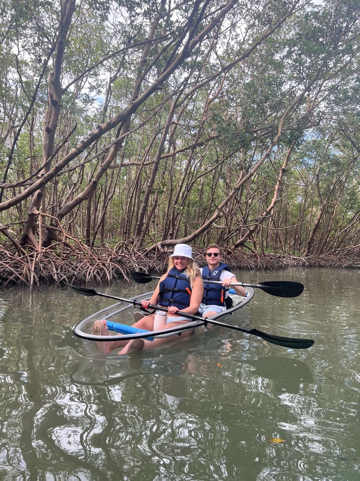 Two paddlers in life vests glide through a mangrove tunnel in a clear-bottom tandem kayak on calm water