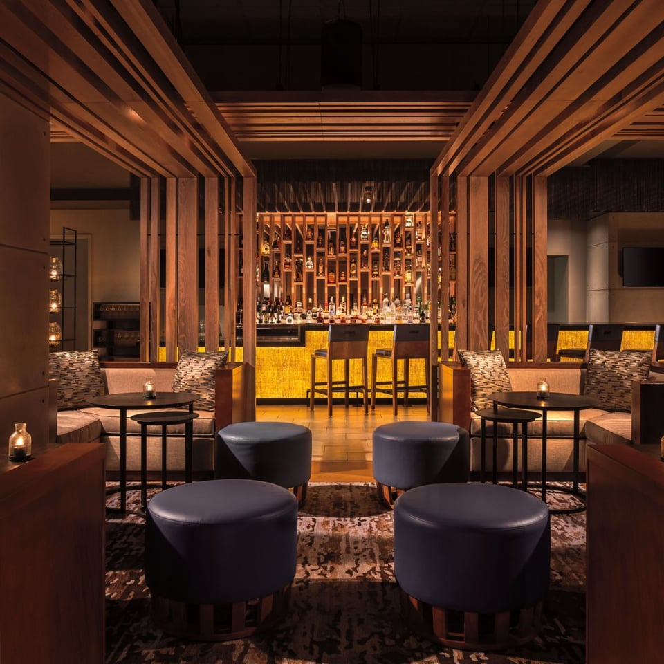 Dimly lit lounge at DUSK with leather ottomans, wood slat partitions and a glowing backlit bar