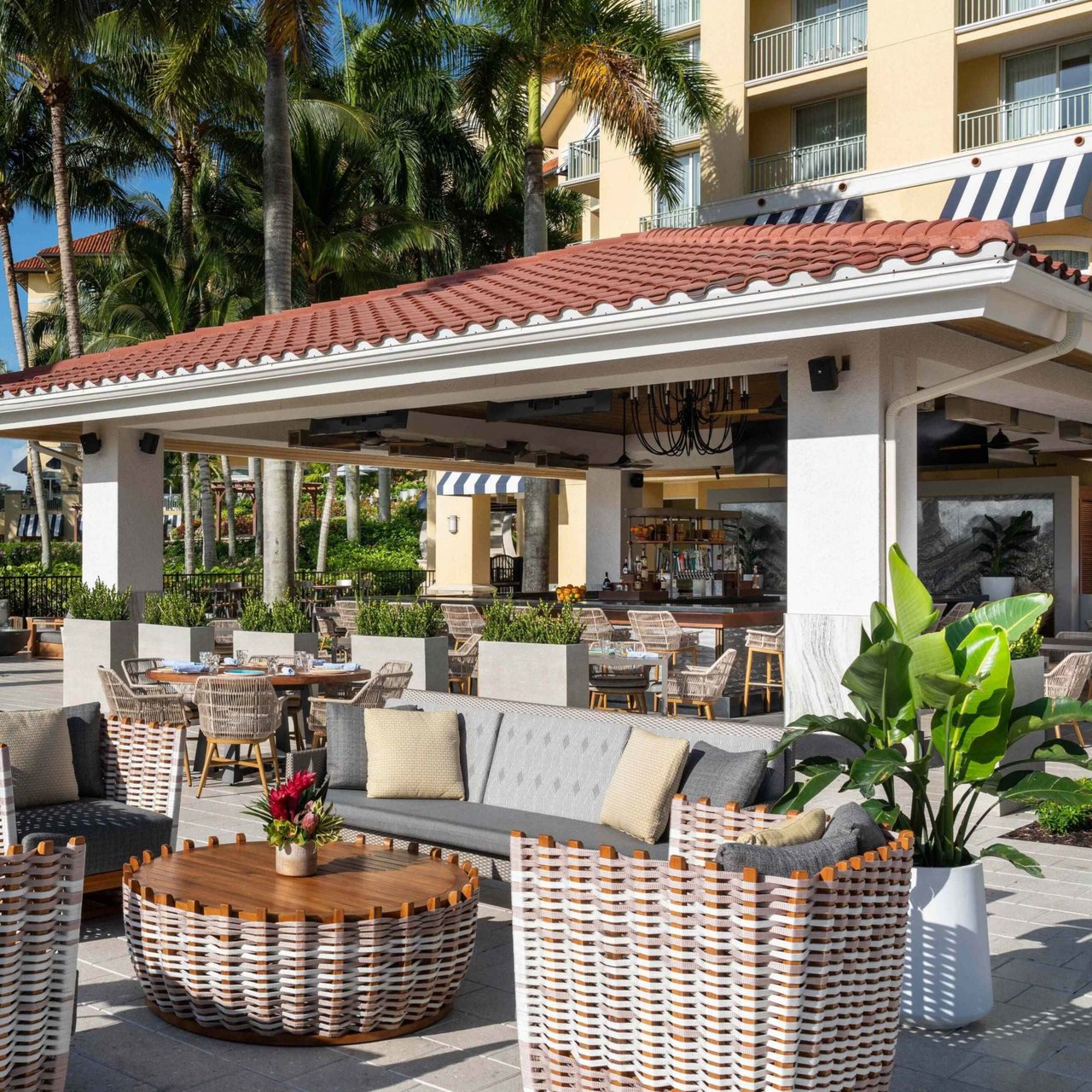 Outdoor patio lounge at Ría with woven armchairs, a tiled-roof bar and palm trees in the afternoon sun