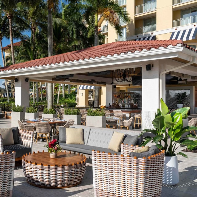 Outdoor patio lounge at Ría with woven armchairs, a tiled-roof bar and palm trees in the afternoon sun