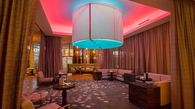 Modern hotel lounge with an oversized drum pendant light, pink and teal ceiling glow, curtains and banquette seating