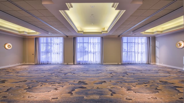 Empty hotel ballroom with patterned carpet, tiered coved ceilings and tall curtained windows glowing with daylight