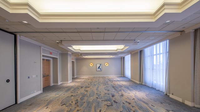 Empty hotel ballroom with patterned carpet, tray ceilings, sheer-curtained windows and wall sconces flanking framed art