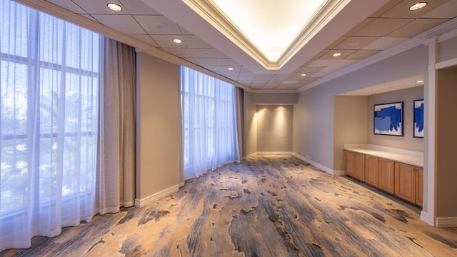 Empty hotel event room with sheer-curtained windows, patterned carpet, recessed lighting and a wood service counter