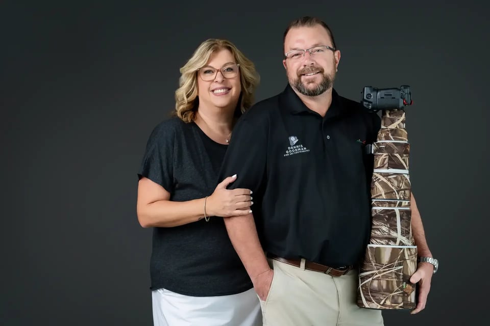 Smiling couple posing against a dark backdrop with a camoflauge-wrapped telephoto camera lens at Dennis Goodman Fine Art Photography