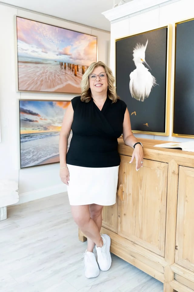 Woman in a black top and white skirt leaning against a wood cabinet in a gallery of beach and egret prints