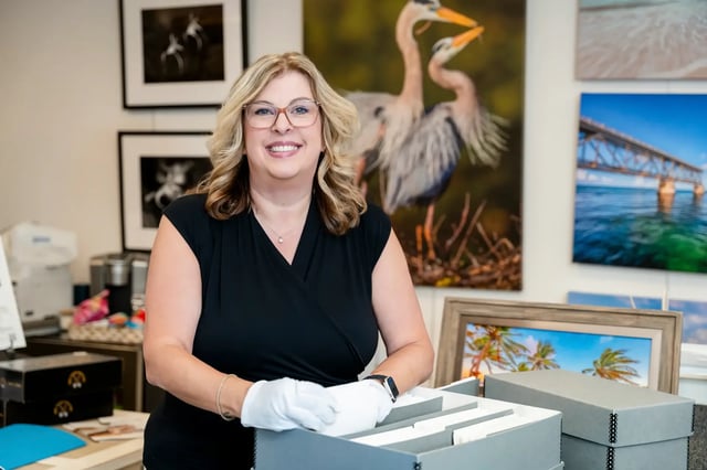 Smiling woman in white handling gloves at a gallery counter with framed heron and beach photographs behind her