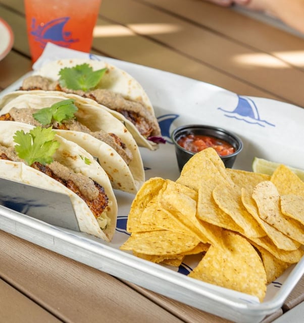 Three soft tacos topped with cilantro served with tortilla chips and salsa on a metal tray at a sunny outdoor table