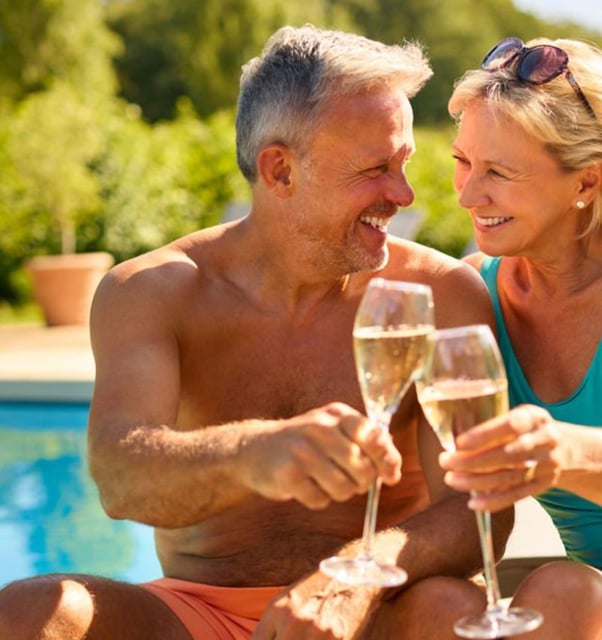 Smiling couple in swimwear toasting champagne flutes beside a sunlit pool