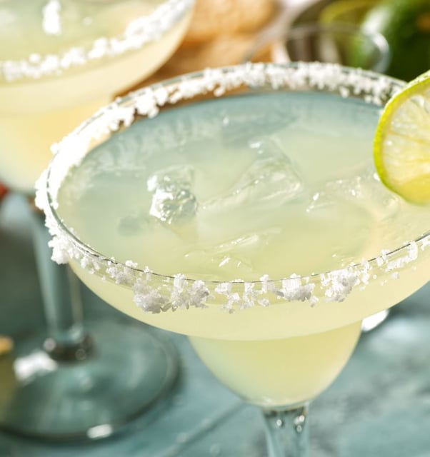 Two salt-rimmed margaritas on the rocks with a lime wheel garnish on a pale blue table