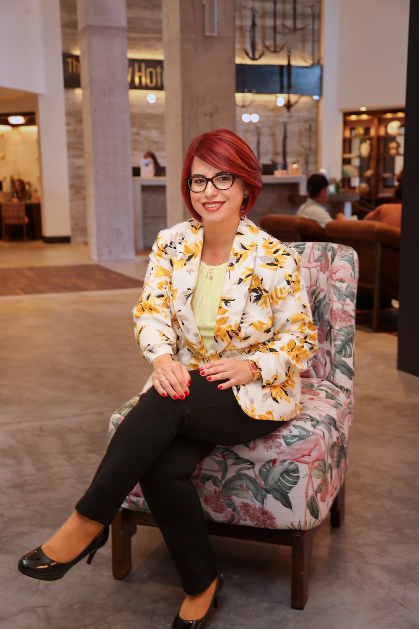 Woman with red hair and glasses in a floral blazer seated in a botanical-print armchair in a hotel lobby