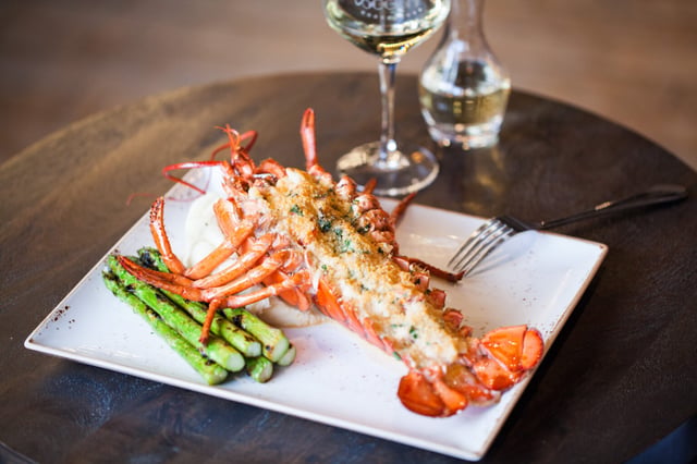 Whole split lobster with breadcrumb topping and grilled asparagus on a square white plate beside white wine