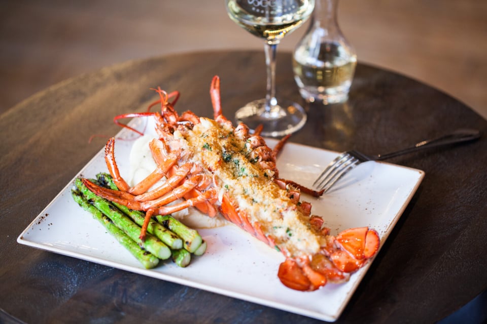 Whole split lobster with breadcrumb topping and grilled asparagus on a square white plate beside white wine