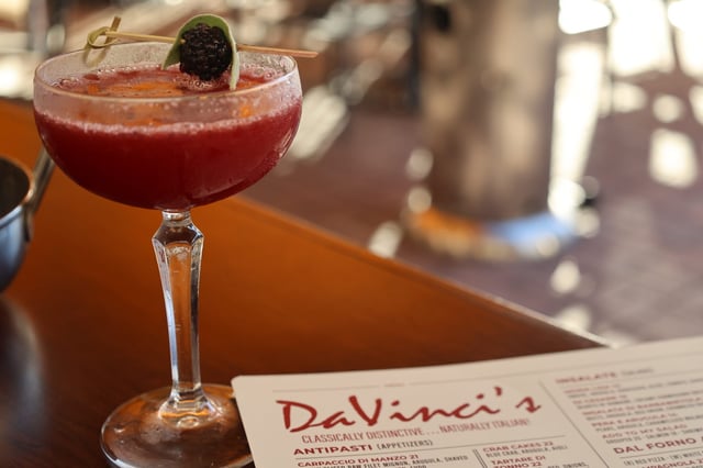 A deep red cocktail garnished with blackberry and lime on a wooden bar beside a DaVinci's menu