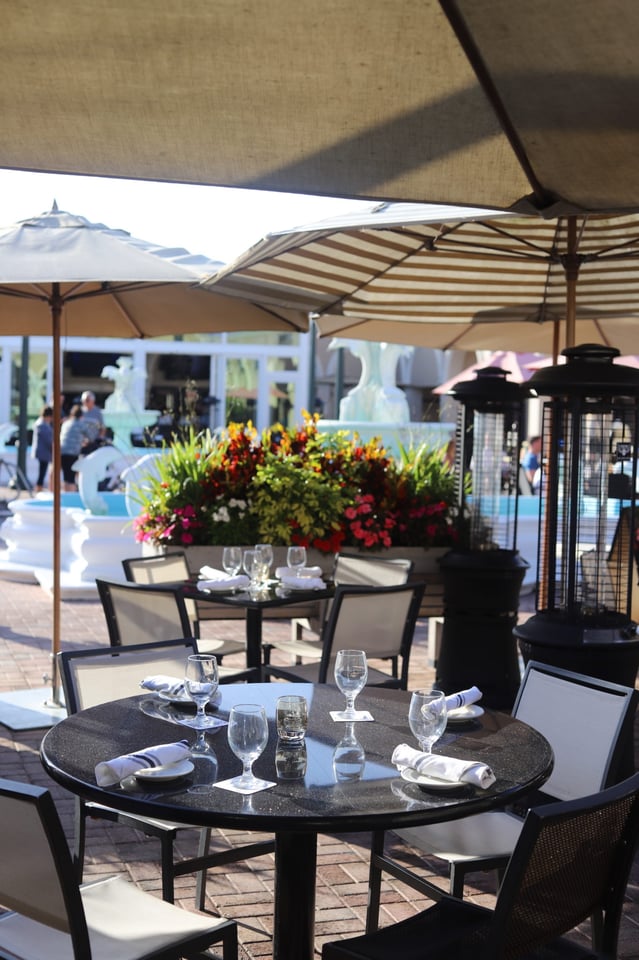 Outdoor patio tables set with wine glasses and napkins under striped umbrellas beside a flower-filled fountain at DaVinci's