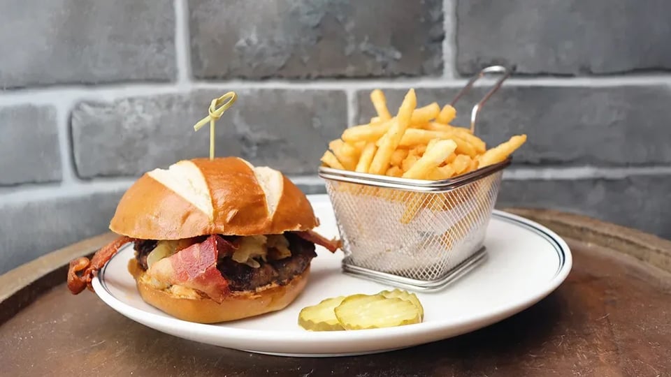 Bacon burger on a pretzel bun with pickles and a wire basket of fries on a white plate at 10K Alley