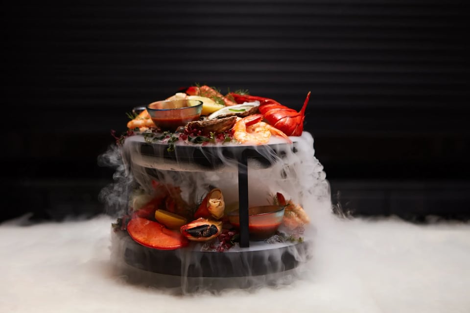 Two-tiered seafood tower with lobster, shrimp, mussels and stone crab claws surrounded by dry ice fog at Ario