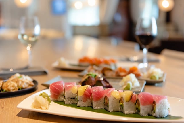A tuna-topped sushi roll on a white platter with wine glasses and more plates blurred behind at Korals Sushi & Cocktail Lobby Bar
